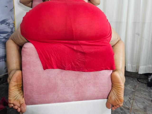 De enorme, rode, doorschijnende billen van NaomyHott69 zitten op een roze stoel, waarbij haar dikke, bruine benen en de zolen van haar voeten in de voorgrond zichtbaar zijn..
