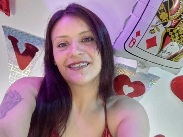 AriannaSeete with long brown hair, smiling, showing metal teeth, wearing a red halter top, heart tattoos on arm, card-shaped decoration, and pinkish-purple light.