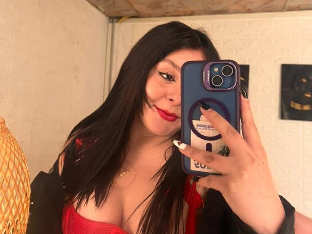 EmaJames, con cabello negro largo, lápiz labial rojo y un sostén rojo que deja ver sus senos en un selfie frente al espejo, sosteniendo un teléfono azul con un agarre de anillo..