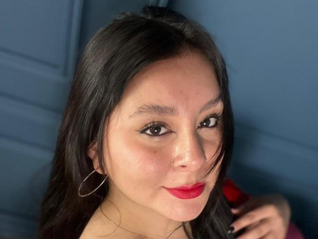 EmaJames has dark brown hair, red lipstick, and black eyeliner. She's wearing hoop earrings and a gold necklace, with a slight smile and a cute, sexy face.