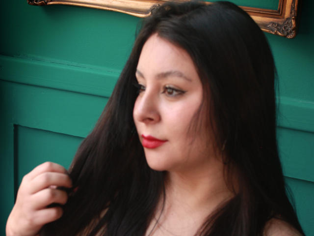EmaJames has long black hair, red lipstick, and a fair skin tone. She's looking to the side with a slight smile, touching her hair with a hand.