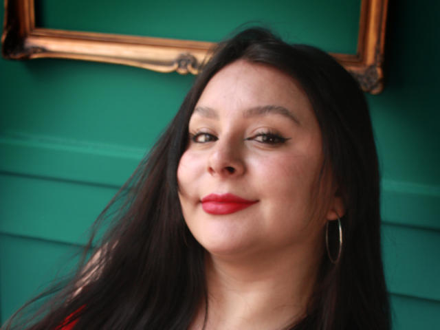 EmaJames has long black hair, red lipstick, and a slight smile, with hoop earrings and a green wall in the background, looking cute and confident.