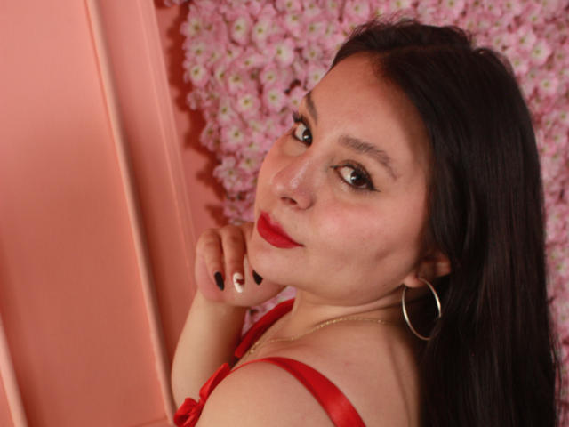 EmaJames with red lipstick, black nail polish, hoop earrings, and red dress, looking over her shoulder, smiling slightly, showing her tits, in a pink-flowered background.