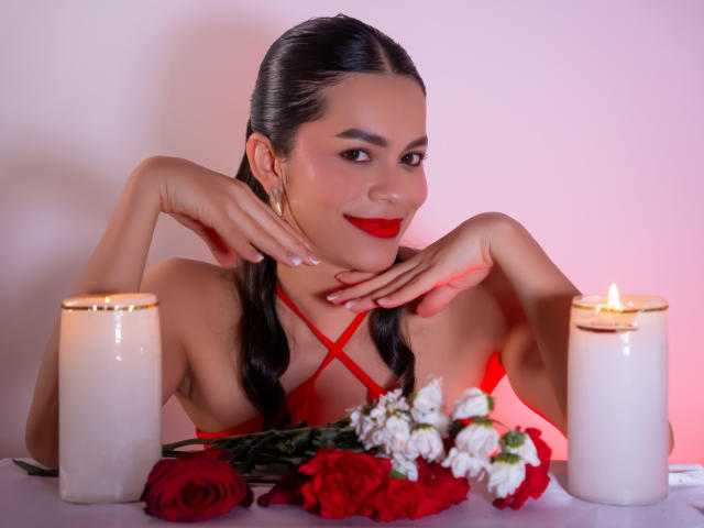 AlanaBlake with red lipstick, black pigtails, and red dress, hands near face, flanked by candles, red and white flowers on table, smiling at camera.