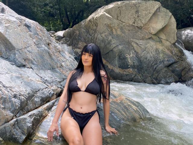 MissValentine stands in a rocky stream, wearing a black bikini. She has long, straight black hair with bangs, and a medium build with a slightly curvy figure. Her bikini has a triangular top and side-tie bottoms. She is leaning on a large rock with her right hand, and the water is flowing around her legs. The rocks are gray and textured. The image is outdoors with greenery in the background. Natural light highlights her medium skin tone.
