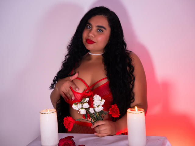 LisaTobon with long black curly hair, red lipstick, wearing red lace lingerie, holding white and red flowers, touching her tits, two lit candles on a white table.