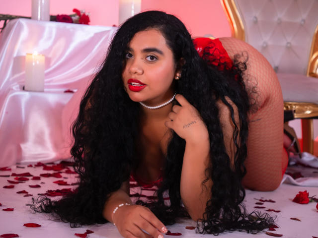 LisaTobon on all fours, wearing red lingerie, fishnet stockings, and a pearl necklace, with long curly black hair, red lipstick, and a tattoo on her right hand. Boobs visible.