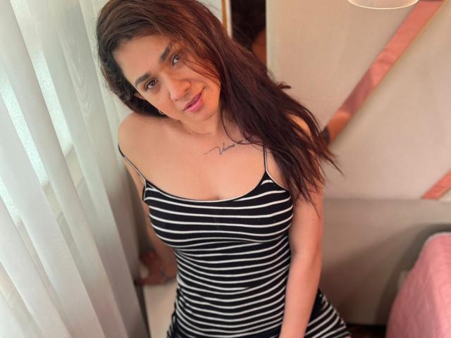 AmmandaMilf with long brown hair, wearing a black and white striped dress, showing her small tits and a tattoo under her collarbone, smiling at the camera.