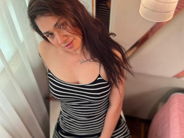 AmmandaMilf with long brown hair, wearing a black and white striped tank top, showing her big tits, sitting near a window, smiling, and having a tattoo on her chest.