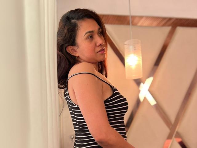AmmandaMilf with dark wavy hair, brown eyes, and a medium build, wearing a black and white striped tank top, standing in a softly lit room, looking over her shoulder.