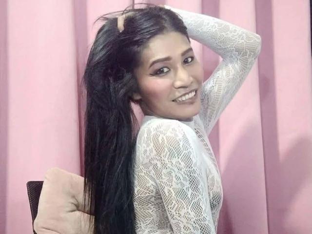 MisstessGalaxia-hot with long black hair, wearing a white lace top, smiling, hand in hair, pink background, cute, pretty, small tits, soft makeup, pink pillow on left.