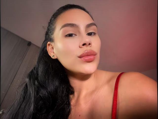 RossaHot with pouty pink lips, dark eyebrows, and long black hair, wearing a red strap, has a cute face, small nose, and is showing off her ears.