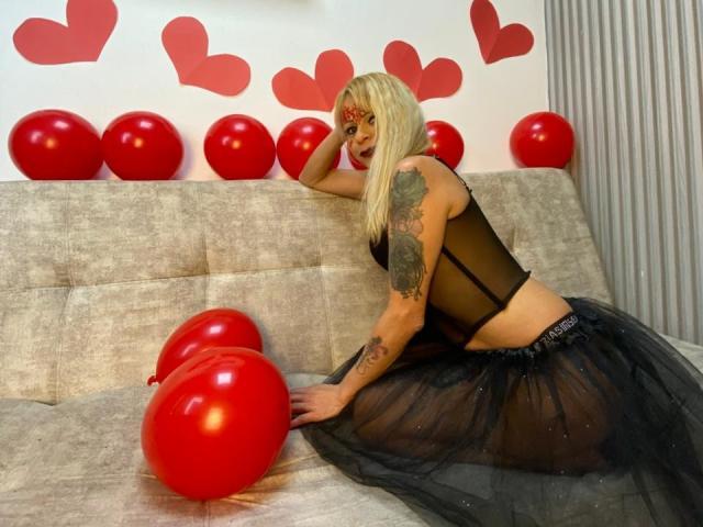 SamanthaJoneX with blonde hair, black sheer top, and black tulle skirt, tattooed arm, red balloons, and hearts, on a grey couch, looking over shoulder, ass slightly visible.