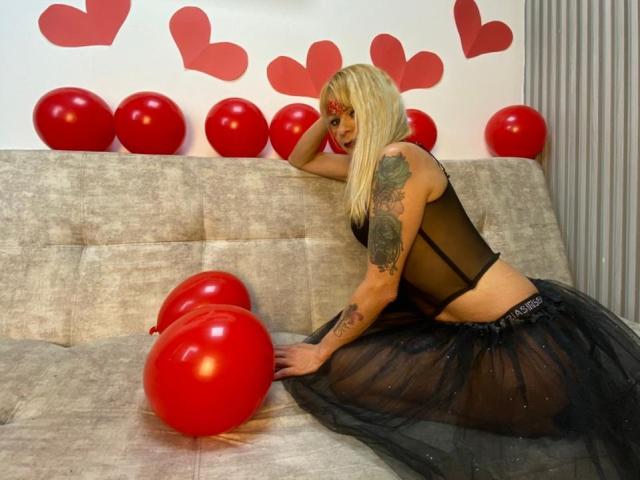 SamanthaJoneX with blonde hair, black sheer top, and black tulle skirt, kneeling on gray couch, red balloons, tattooed arm, and red heart decals in background.