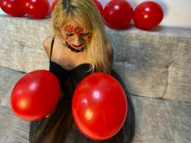 SamanthaJoneX with blonde hair, red face paint, black dress, and red balloons, holding two balloons in front of her, sitting on a gray couch. Boobs, tits, ass not visible.