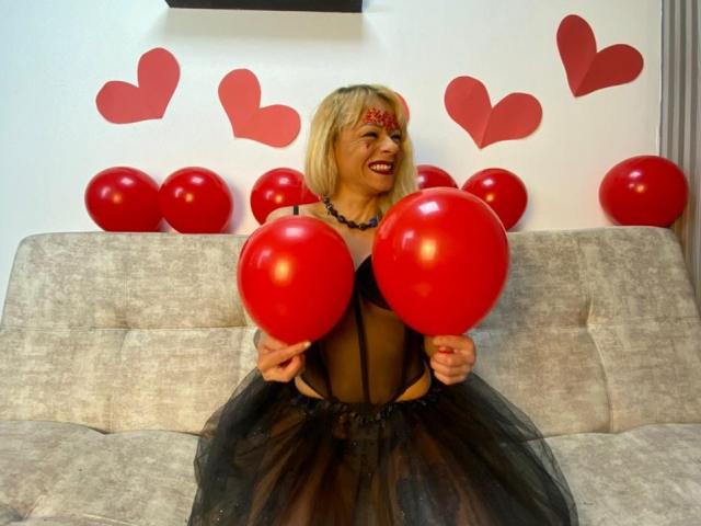 SamanthaJoneX with blonde hair, red face paint, black sheer dress, holding two red balloons, sitting on grey sofa, smiling, surrounded by red balloons and hearts on white wall.