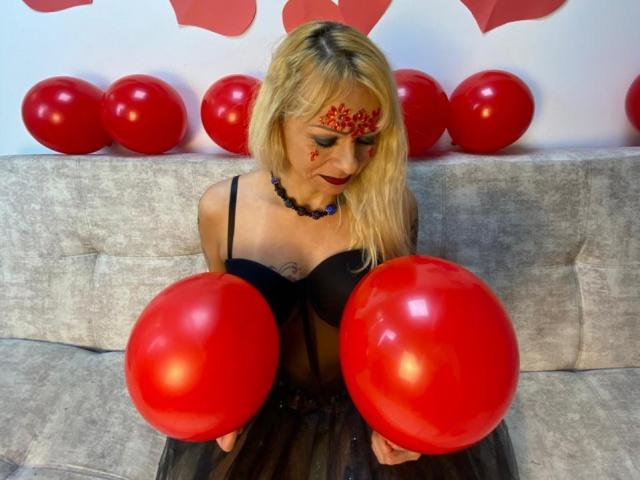 SamanthaJoneX with blonde hair, red face paint, black dress, holding red balloons, sitting on gray couch, black necklace, tattoo on chest, looking down, cute, sexy.