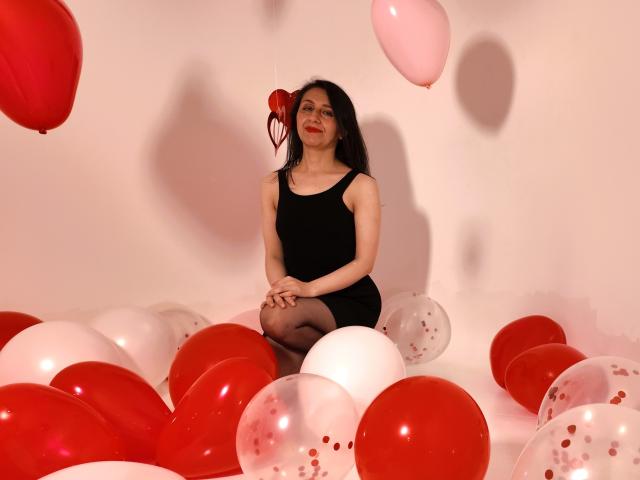 Cyntiia with black dress and black tights, sitting on red and white balloons, red lips, long black hair, and a red heart balloon near her head.