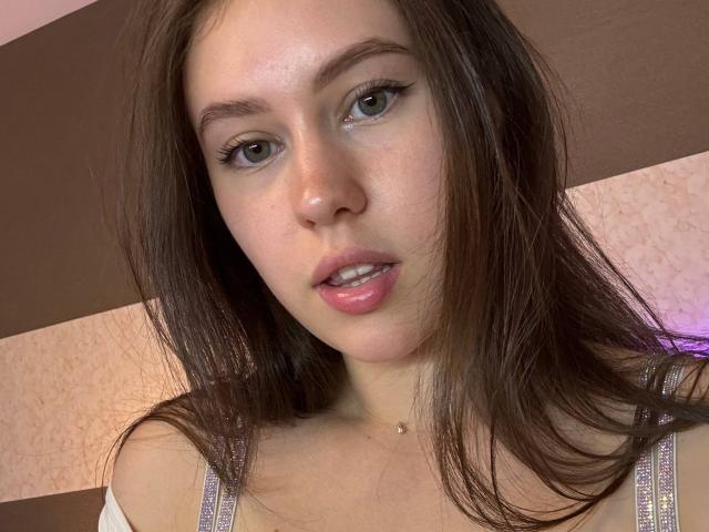Eyira has light brown eyes, straight brown hair, and pink lips. She's wearing a white top with purple straps, and a small necklace. Her tits are partially visible.