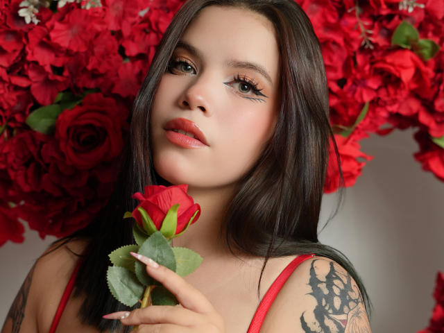 AryButty with long dark hair, red lipstick, holding a red rose, tattoo on right arm, red dress, fluffy red rose petals in background, looking up with seductive eyes.