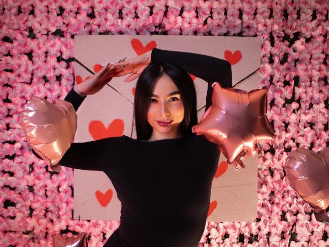 CamilleRios with dark hair, black long-sleeve top, pink heart balloons, and rose-pink star balloons, arms raised, smiling, standing in front of a pink rose wall.