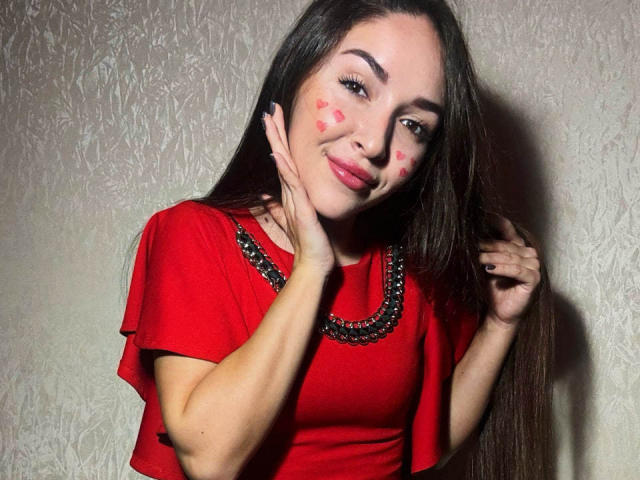 AquaLady with long brown hair, red dress, and black beaded necklace, smiling, hands on cheeks, red heart stickers on her face, looking cute and sexy.
