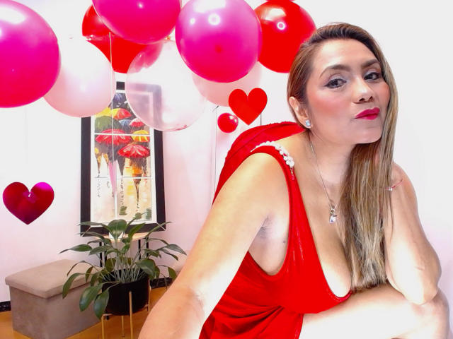 LarisMilfHot with blonde hair, red dress, and large tits, leaning forward, red balloons, pink and white balloons, heart balloons, potted plant, and beige cushion in the background.