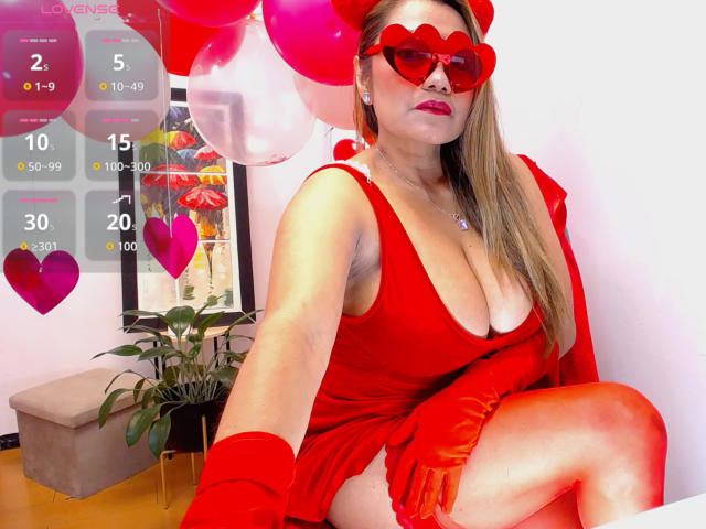 LarisMilfHot with big boobs, wearing a red heart-shaped mask, red dress, and red gloves, sitting with one leg up, surrounded by red and pink balloons.