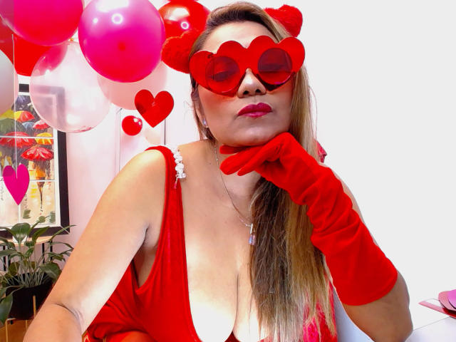 LarisMilfHot with red heart-shaped glasses, red gloves, and red dress, showing her tits, posing with hand on chin, surrounded by pink and red balloons.