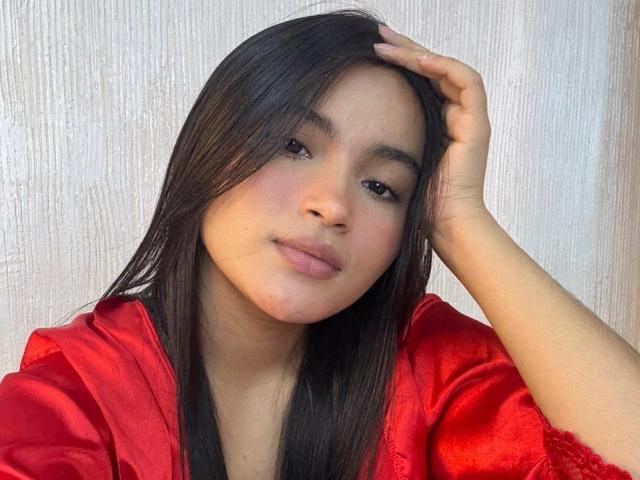 MiiaPalmer with long black hair, wearing a red satin robe, has her hand in her hair, looking at the camera with a subtle smile, soft brown skin, and brown eyes.