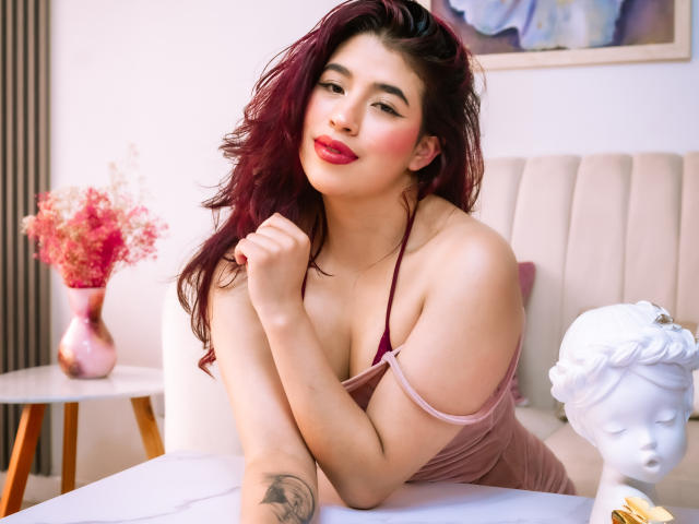 ElizaNior with red lipstick, wavy dark hair, pink dress, showing tits, arm tattoo, white bust, pink vase, smiling, arm on table.