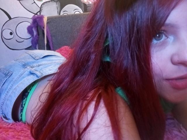 SophieHill with red hair, lying on a pink blanket, wearing green lingerie and denim shorts, showing her ass and a side view of her face.