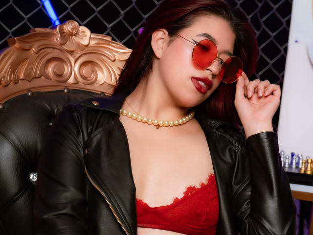 ElizaNior, a young Asian woman with fair skin and dark red hair, wears red lace lingerie, a black leather jacket, and round red sunglasses. She has a pearl necklace and red lipstick. Her right hand touches her glasses. The background has a black diamond-patterned wall and a wooden chair with a gold, ornate headrest. The image has a modern, edgy style with bold colors and textures. Her expression is confident and slightly sultry.