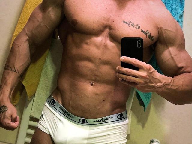 AngeloFitt, muscular, shirtless, white Calvin Klein briefs, holding phone, tattoos on arm and chest, visible abs, flexed arm, towel in background, tan skin, serious expression, mirror selfie.