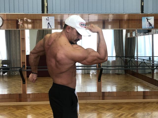 AngeloFitt stands in a dance studio, flexing his right arm. He's muscular with a broad back and defined muscles. He's shirtless, wearing black shorts and a white cap with red text. His skin is tan, and he has a short beard. The wooden floor has a herringbone pattern, and a mirrored wall behind him reflects his pose. The room has large windows with sheer curtains, and a wooden barre is mounted on the wall.