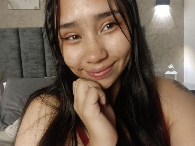 Karollteyes with long black hair, freckled face, brown eyes, and a slight smile, resting her chin on her hand, wearing a red tank top, showing a small tattoo.