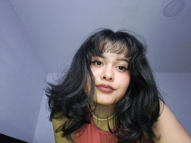 JennLunaLee has black, wavy hair, medium skin, and is wearing a red sleeveless top with a pearl necklace. Her small tits are slightly visible. She's looking down.