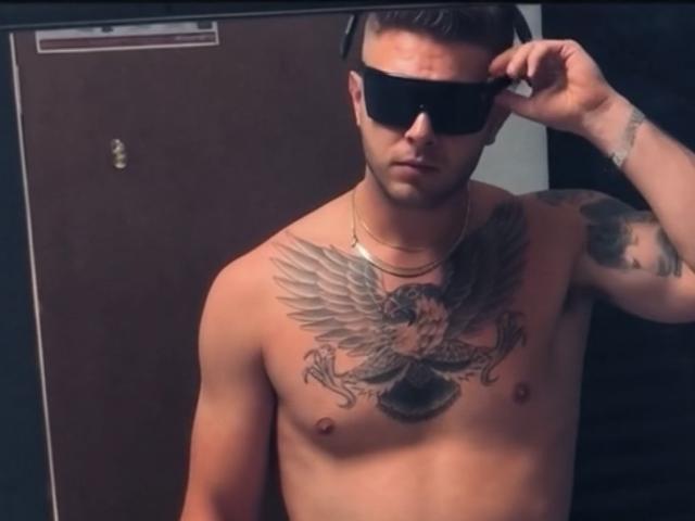 Antzo stands shirtless, wearing black sunglasses, adjusting them with his right hand. He has a muscular build and a large, detailed tattoo of a bird with outstretched wings on his chest. His left arm, partially visible, has more tattoos. Antzo's skin is light, and he has short brown hair. He wears a gold chain necklace. The image is a photograph with a focus on his upper body. Slightly angled, Antzo's serious expression adds a sense of intensity. The background is a dark, plain door.