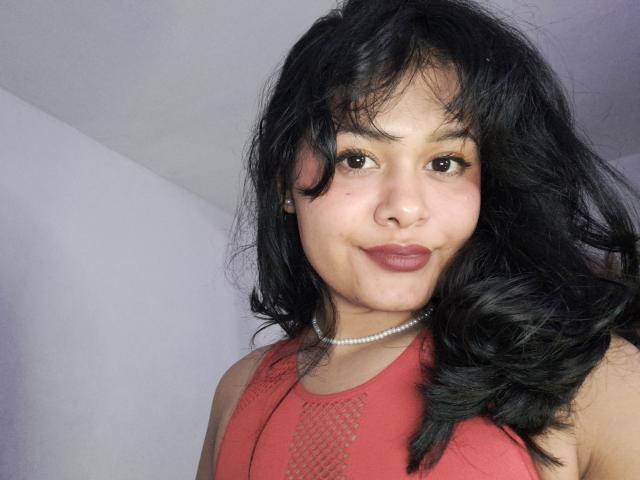 JennLunaLee has medium-length, wavy black hair that partially covers her right shoulder. She has medium brown skin and dark brown eyes. She's wearing a red sleeveless top with a mesh pattern on the chest, and a pearl necklace. Her lips are slightly puckered in a subtle smile. The lighting is even, highlighting her natural skin texture. The image focuses on her upper body, showing a close-up from the chest up.