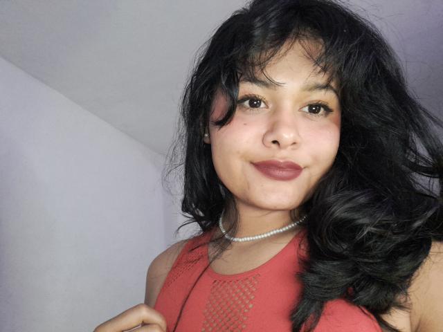 JennLunaLee with black wavy hair, brown eyes, and red lipstick, wearing a red mesh top and pearl necklace, smiling slightly, showing a cute face and small tits.
