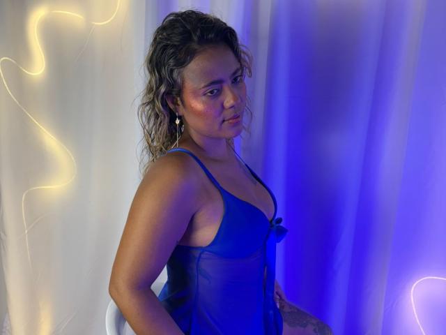 NatachaLatinHot with wavy brown hair, blue sheer lingerie, and visible ass, looking sideways with a subtle smile, glowing yellow and blue lights in the background.