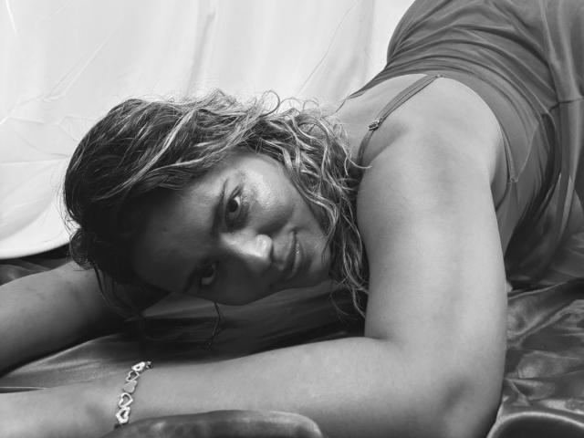 NatachaLatinHot has wavy, wet-looking hair, lying on a shiny surface in a black-and-white shot, wearing a sleeveless top, with a bracelet on her right wrist, looking at camera.