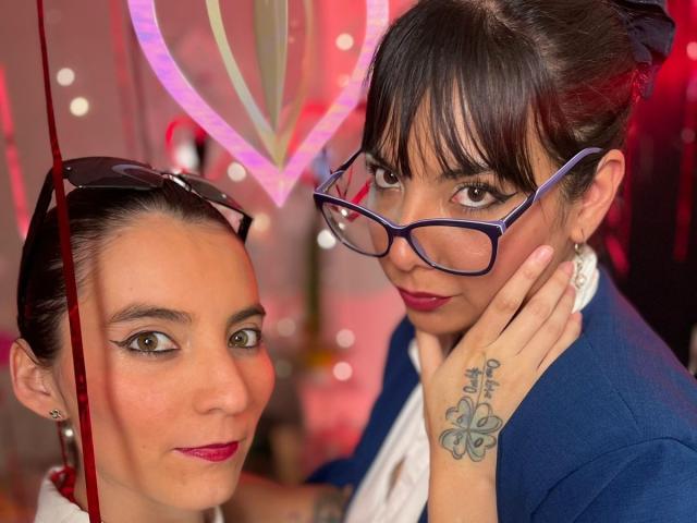 ValkiriaAndDione with glasses, blue blazer, white shirt, and tattoo on hand, touching face of ValkiriaAndDione with red lipstick, brown hair, and sunglasses on head. Boobs are not visible.