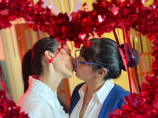Two ValkiriaAndDiones kissing, facing each other, with red tinsel and ribbons in the foreground. The ValkiriaAndDione on the left has dark hair in a ponytail, wearing a white shirt. The ValkiriaAndDione on the right has dark hair in a bun, wearing blue glasses and a blue jacket over a white shirt. Warm yellow background. Sharp contrast between red tinsel and ValkiriaAndDiones. Intimate, close-up shot.