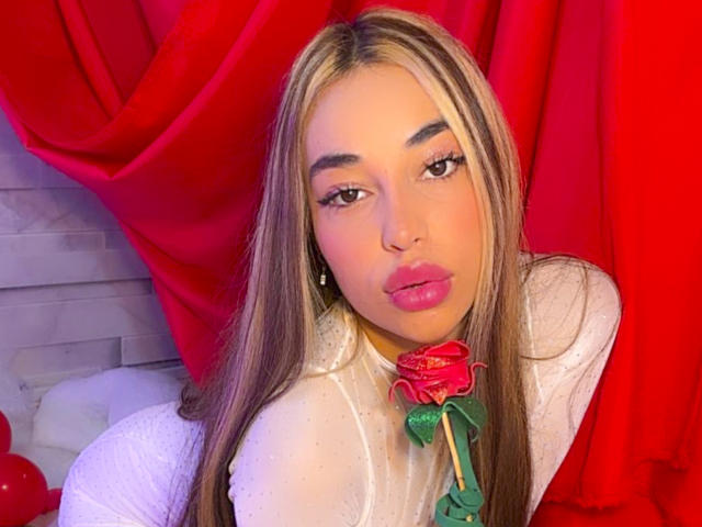 MissVallen with long blonde and brown hair, wearing a white sheer top, holding a red rose, pouting with pink lips, red curtain in background.