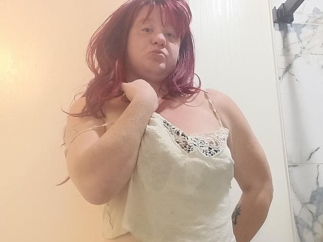 DeliciousRain with red hair, wearing a white lace-trimmed camisole, pouting, left hand near her neck, slightly chubby, visible tattoo on right arm.