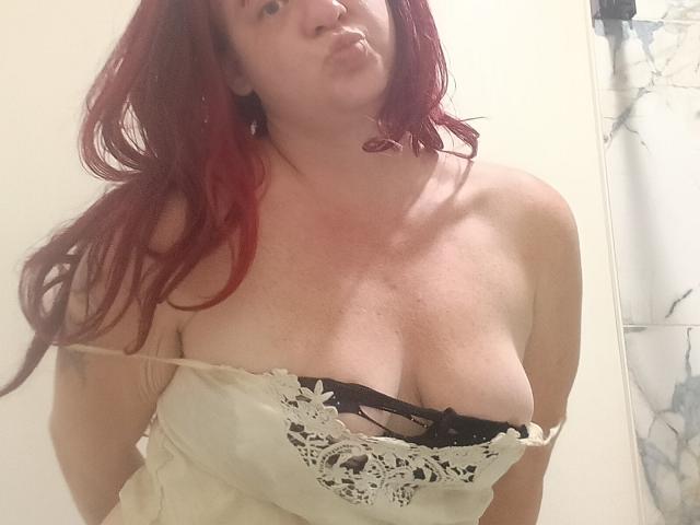DeliciousRain with red hair, puffy lips, fair skin, and large tits, wearing a white lace-trimmed top that exposes black bra and cleavage.
