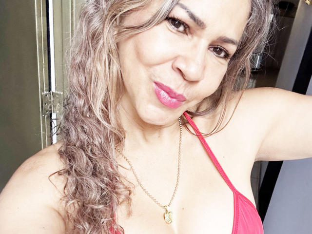 BrigiteWalsh has curly, light brown hair, pink lipstick, and a gold necklace. She's wearing a pink, one-shoulder top, showing her small tits and smiling at the camera.