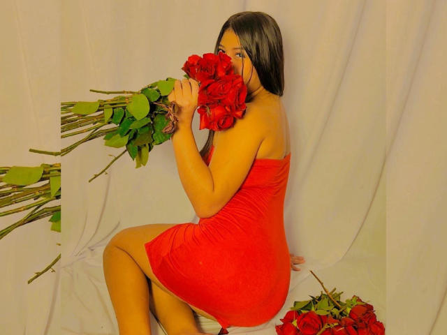 BadKristty with long black hair, wearing a tight red dress, sitting on white fabric, holding red roses over her face, with more roses on the floor and left.