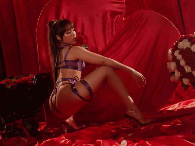 Heyly in purple lingerie, squatting on red satin, holding a red rose, with a bouquet of roses, wearing high heels, her ass and tits prominent, her ponytail back.